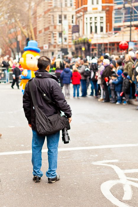 travel photgrapher in a black parka holding a DSLR watching people in a parade at a distance.